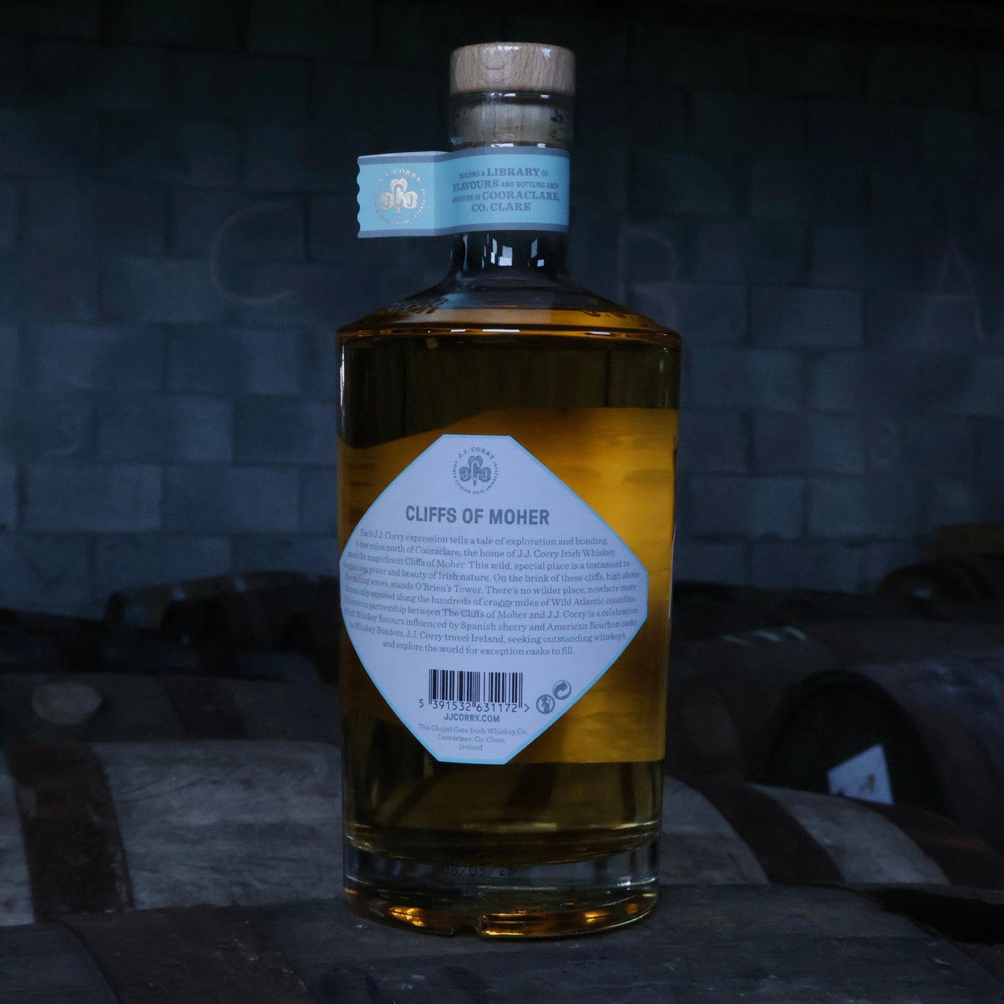 This is a photograph of a bottle of JJ Corry's The Cliffs of Moher Irish whiskey on top of a wooden whiskey cask with a dark background