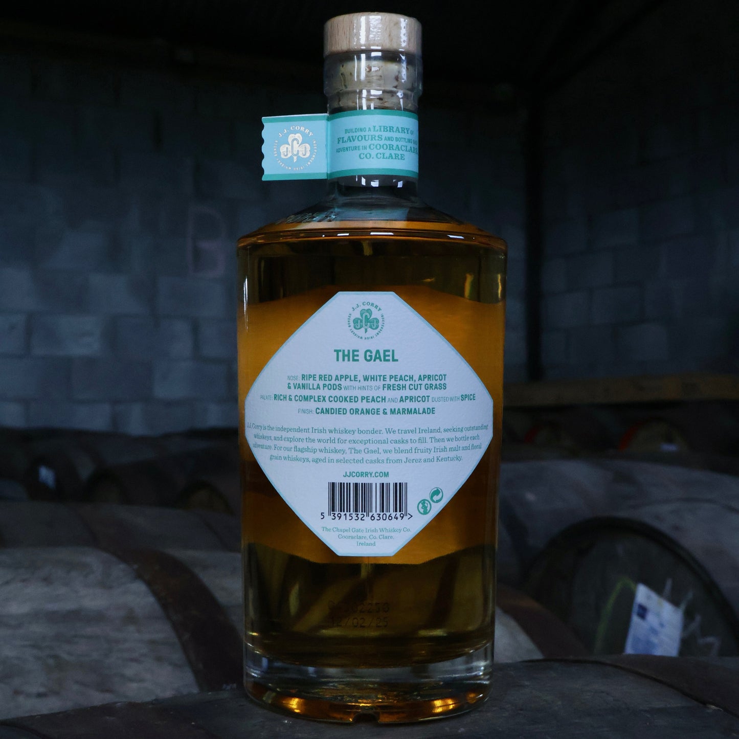 This is a photograph of a bottle of JJ Corry's The Gael Irish whiskey on top of a wooden whiskey cask with a dark background