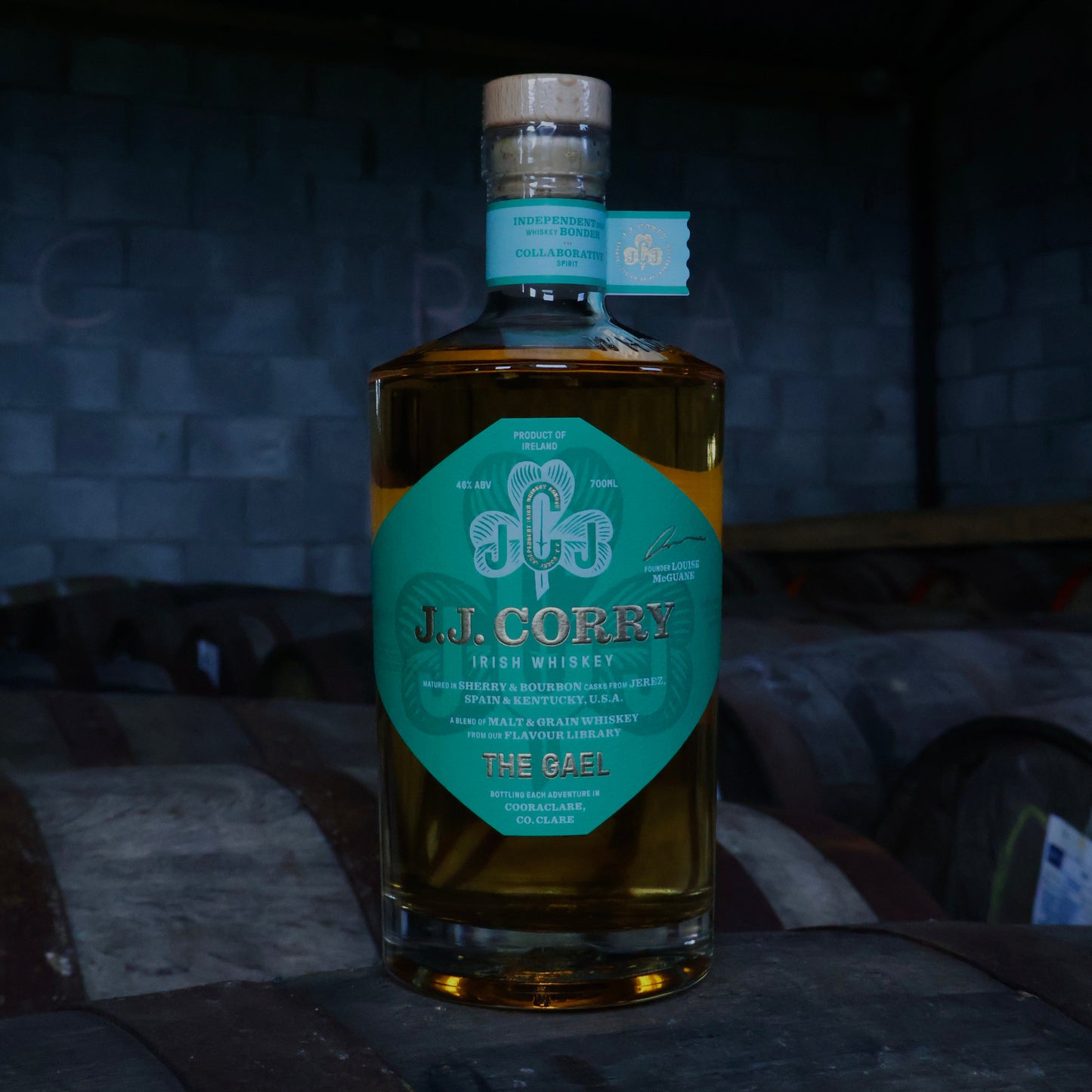 This is a photograph of a bottle of JJ Corry's The Gael Irish whiskey on top of a wooden whiskey cask with a dark background