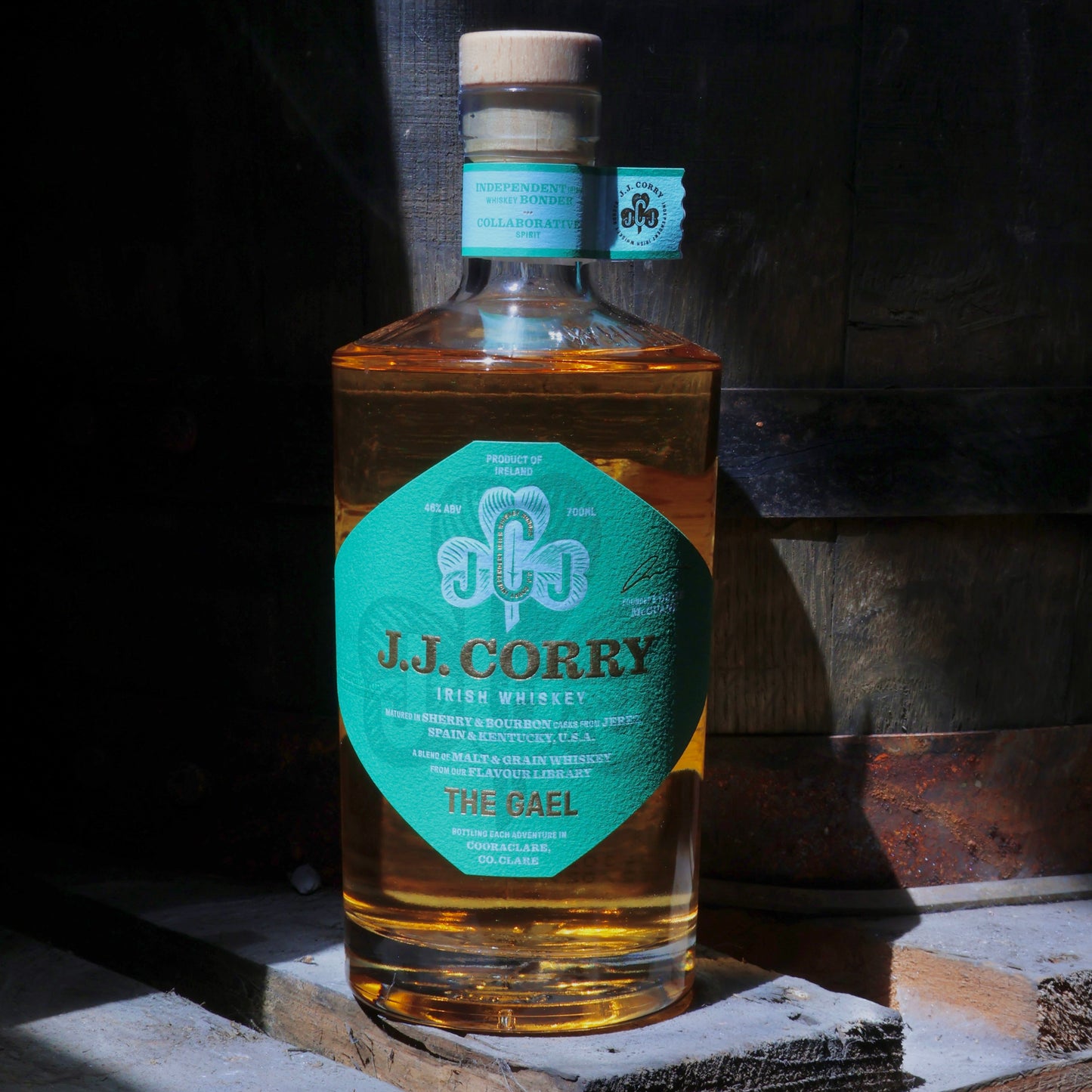 This is a photograph of a bottle of JJ Corry's The Gael Irish whiskey on top of wood with a wooden cask in the dark background