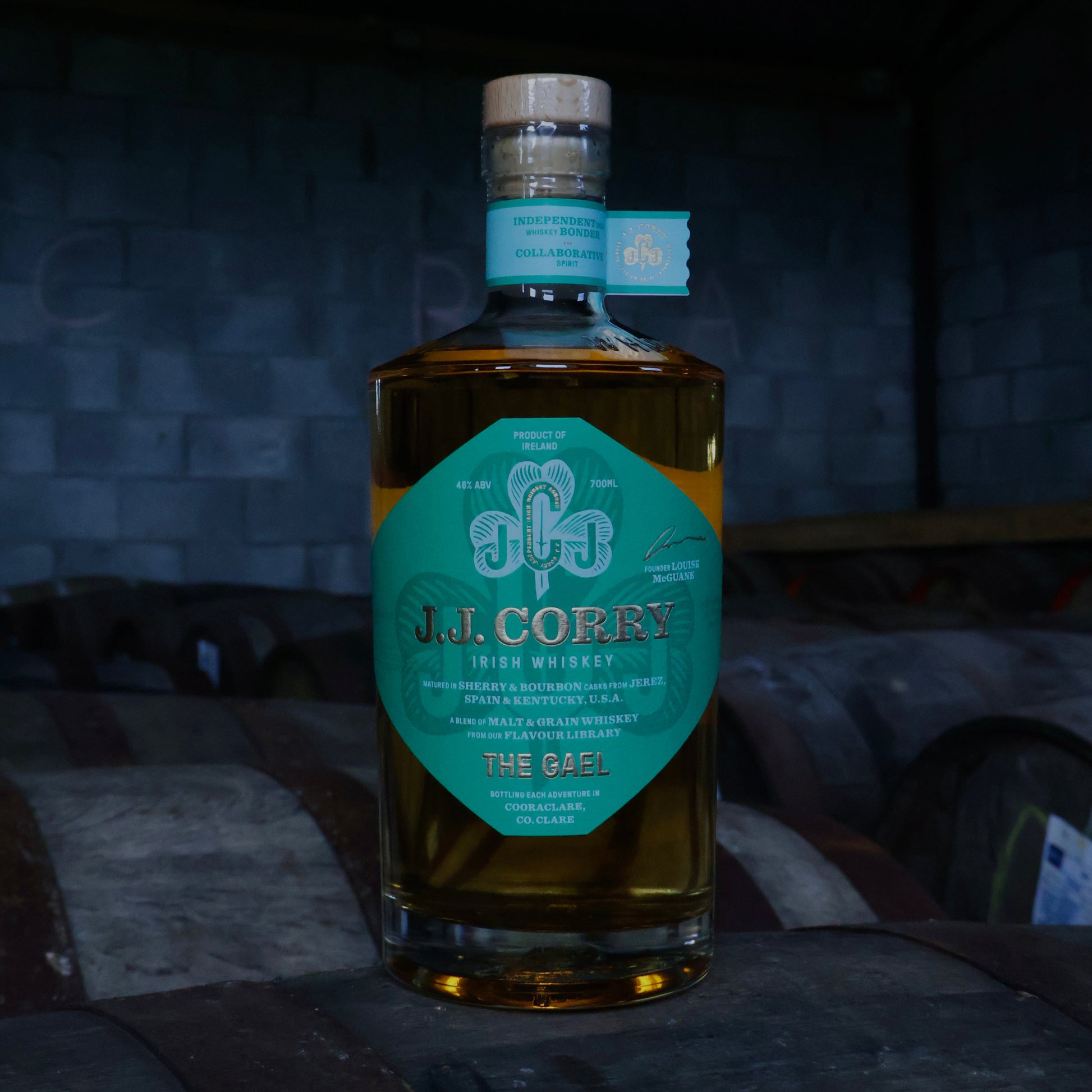 This is a photograph of a bottle of JJ Corry's The Gael Irish whiskey on top of a wooden whiskey cask with a dark background